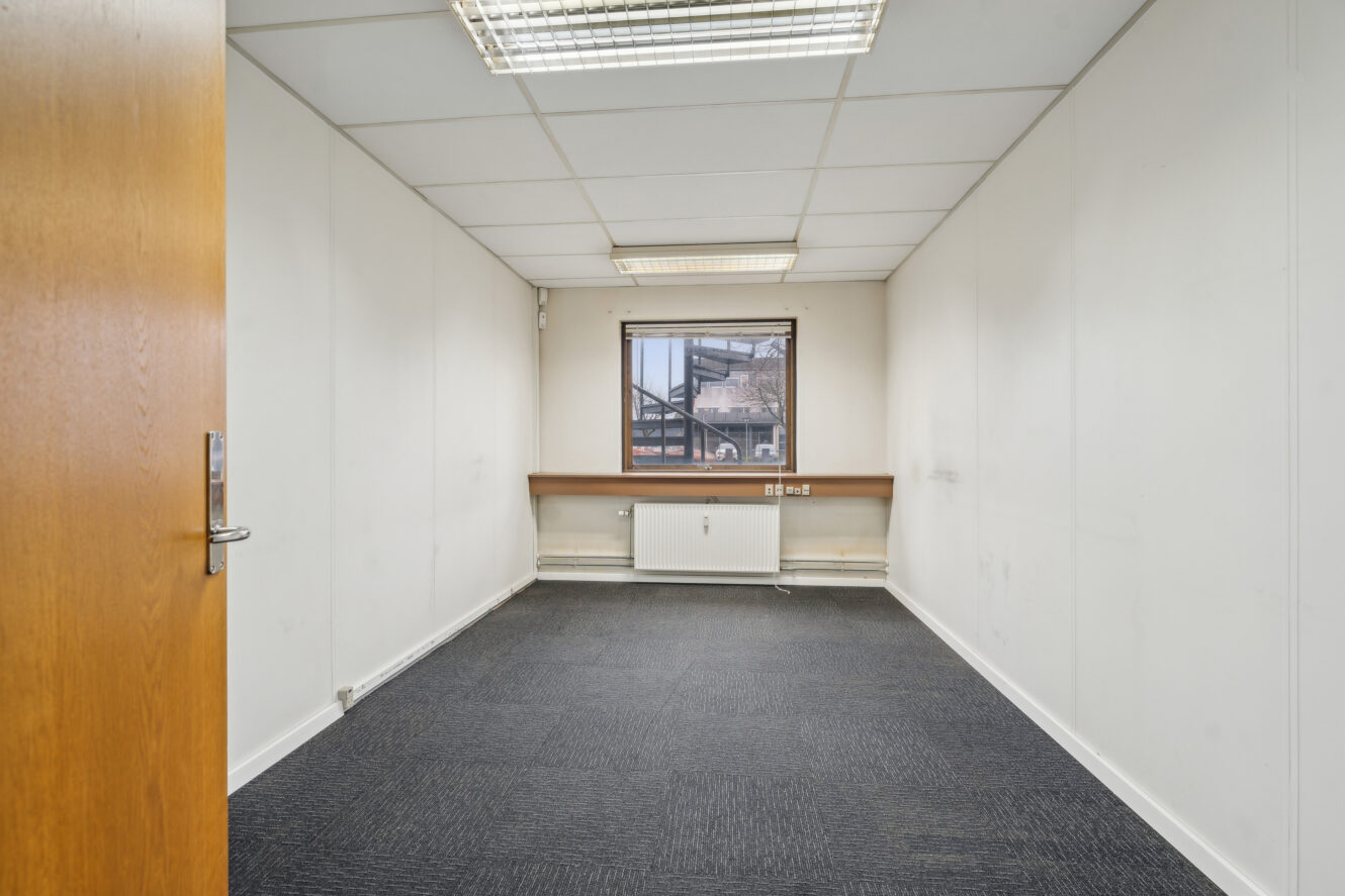 Et lille, tomt kontorlokale med hvide vægge, et vindue, loftslamper, mørkt gulvtæppe og en radiator under vinduet. Døren er delvist åben.