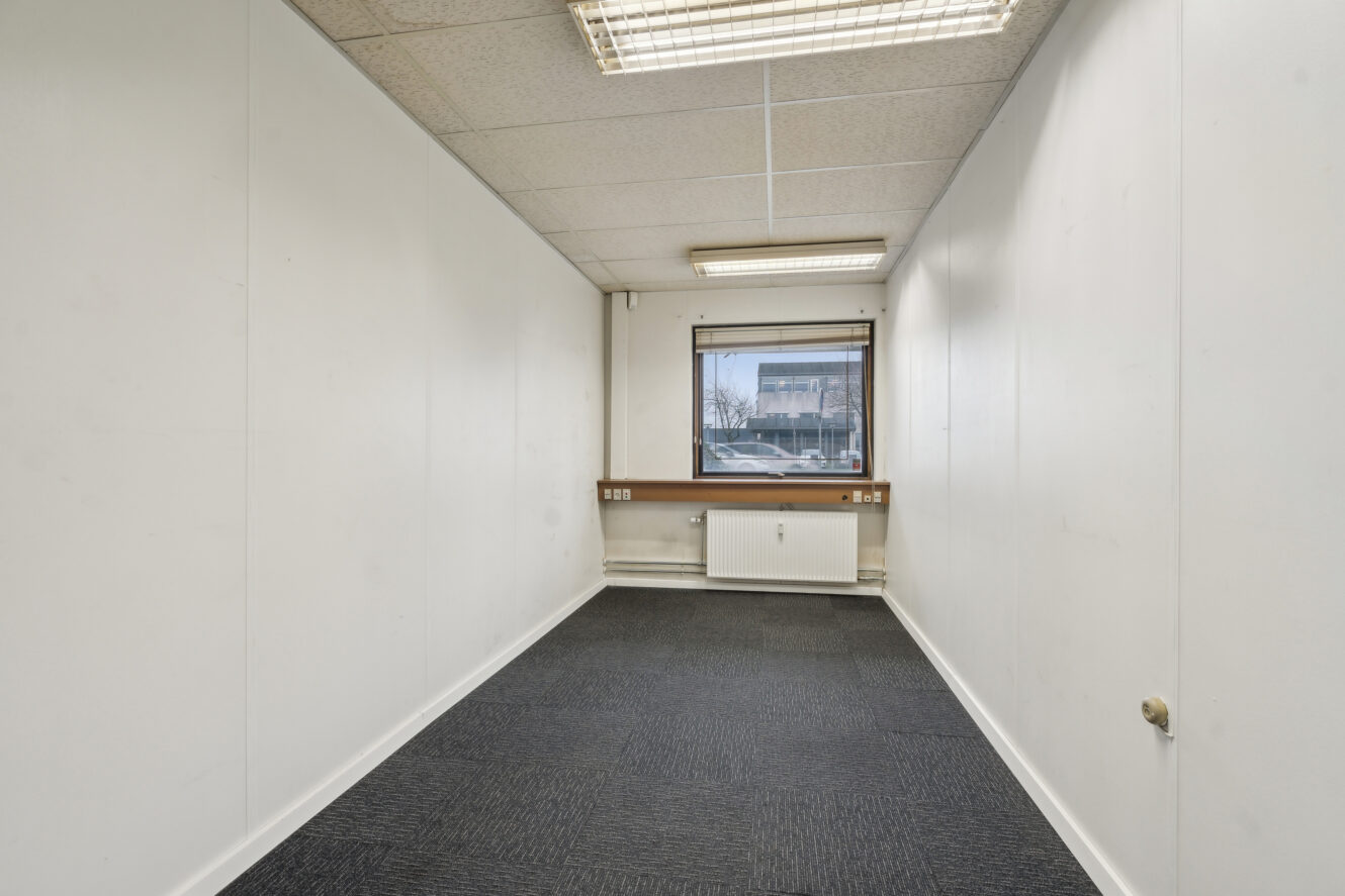 Et lille, tomt kontorlokale med hvide vægge, et mørkt gulvtæppe, et vindue, en radiator og lysstofrør i loftet.