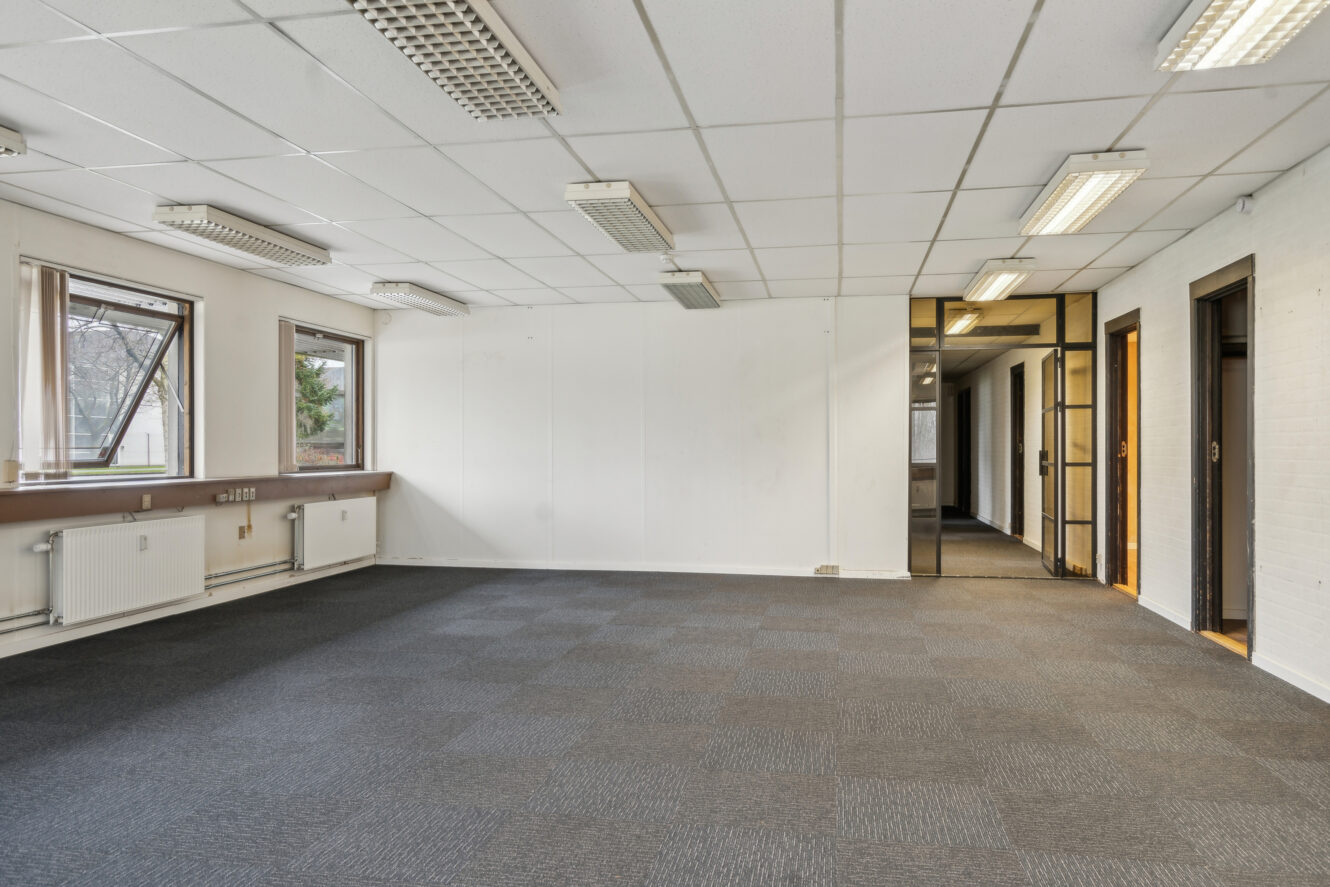 Tomt kontorlokale med gråt gulvtæppe, hvide vægge, lysstofrør i loftet, tre vinduer til venstre og døråbninger, der fører til tilstødende lokaler.