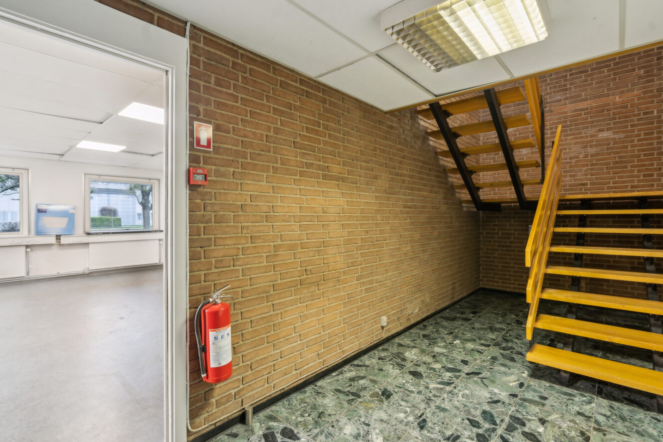 En trappeopgang med gule trin og en murstensvæg med en brandslukker og en alarm; et tilstødende rum med vinduer og radiatorer er synligt gennem en åben dør.