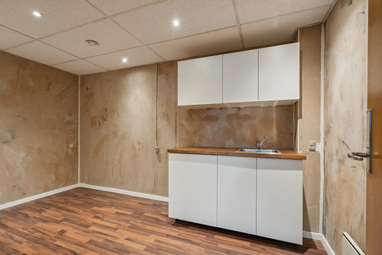 Lille tekøkken med hvide skabe, en vask og træbordplade mod ufærdige gipsvægge. Loftet har indbyggede lamper, og gulvet er mørkt trælaminat.
