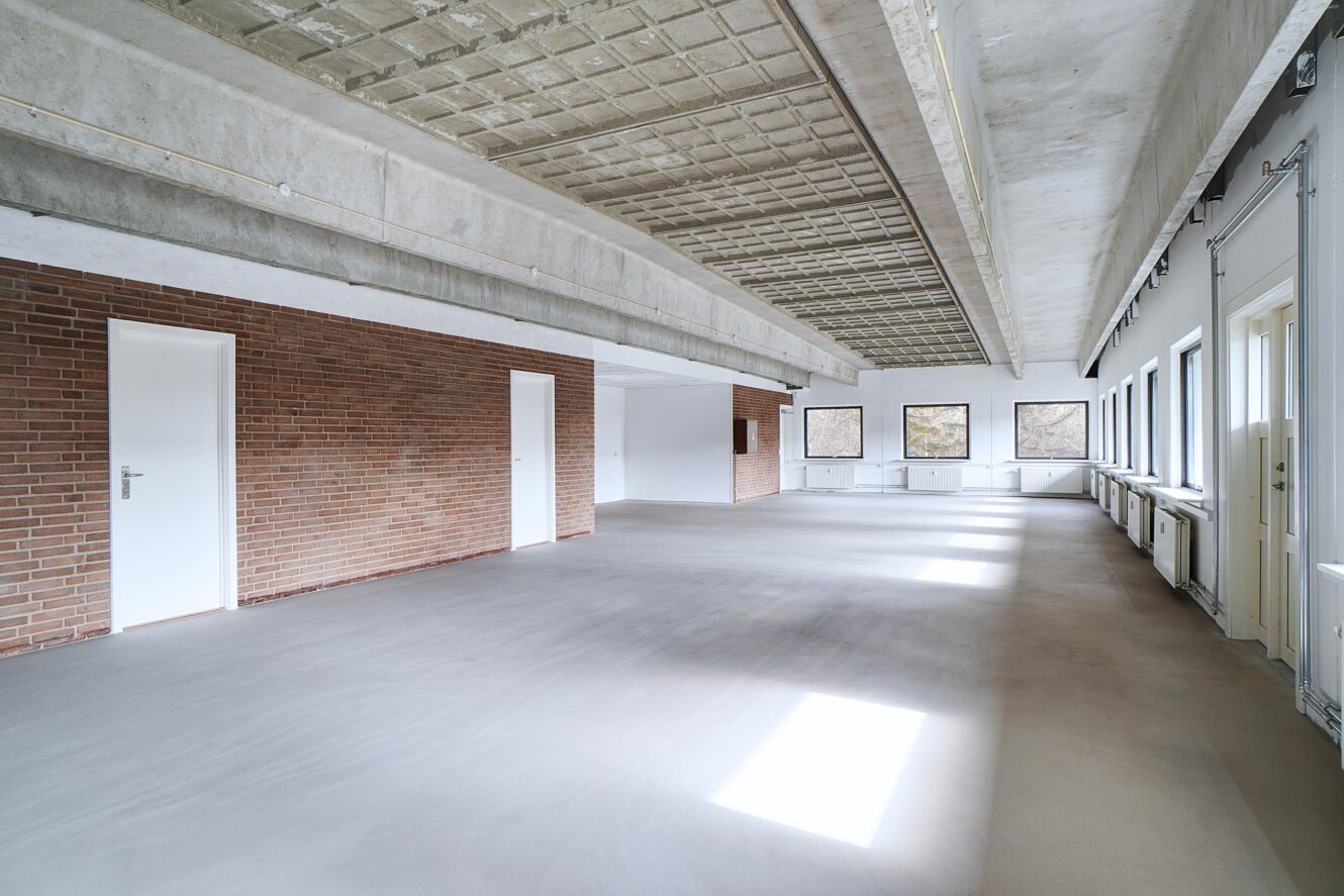 Et stort, tomt rum med betonloft, murstensvægge, flere hvide døre og vinduer, der lukker naturligt lys ind.