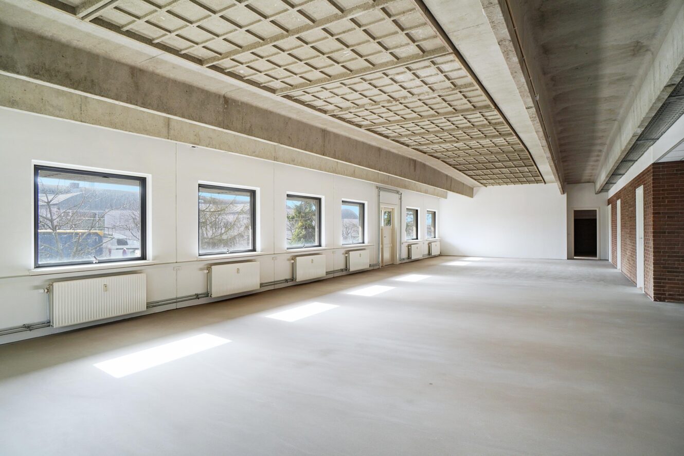Et stort, tomt rum med hvide vægge, flere vinduer, eksponeret betonloft og et bart gulv; naturligt lys kommer ind gennem vinduerne.