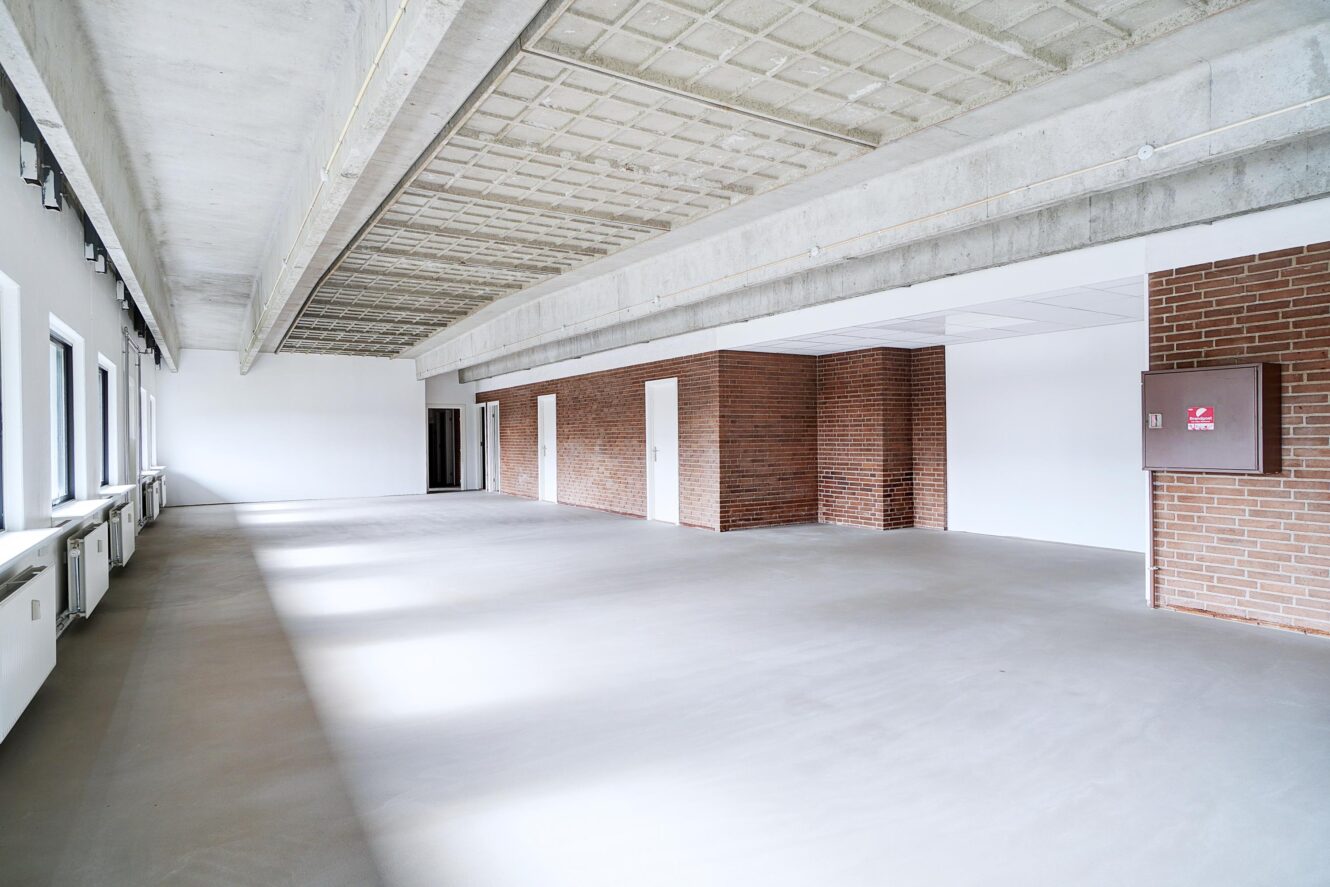Tomt rum i industristil med betongulv, synlige loftsbjælker, indvendige murstensvægge og flere vinduer langs den ene side.