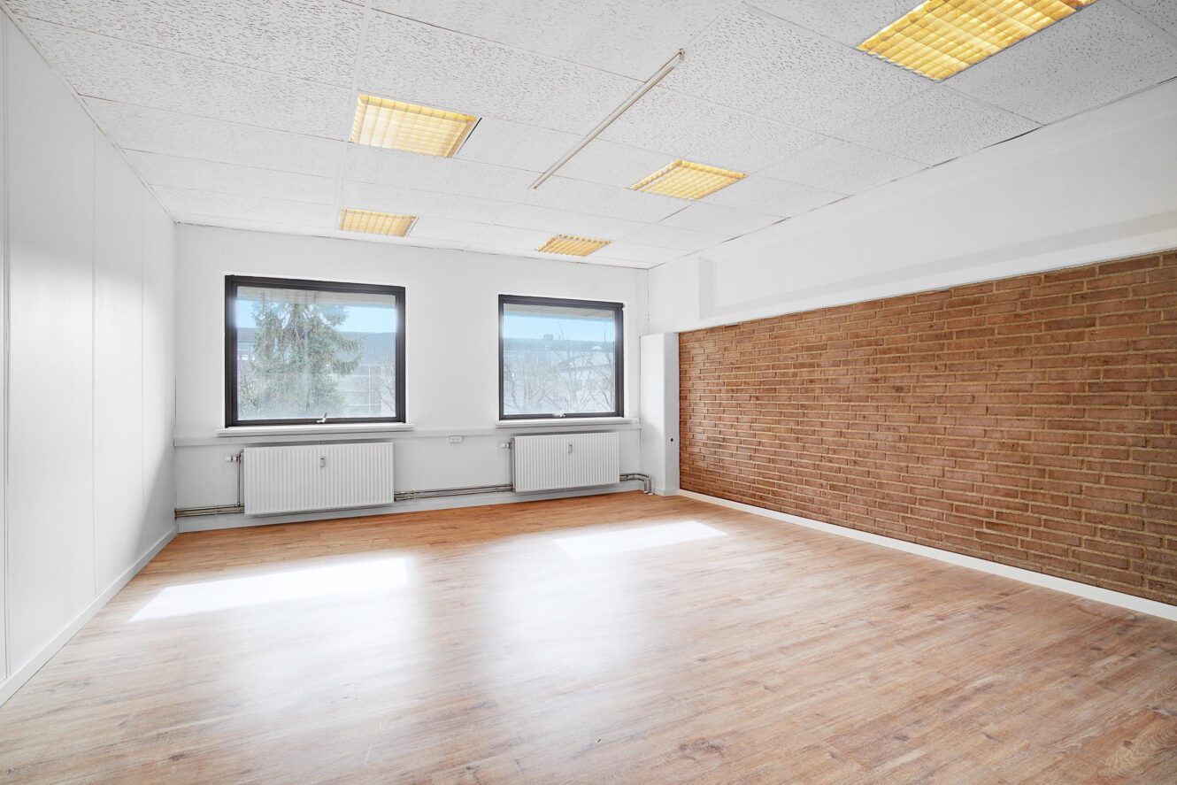 Tomt rum med to store vinduer, en murstensvæg med accent, hvide loftsplader med lysstofrør og trægulv.