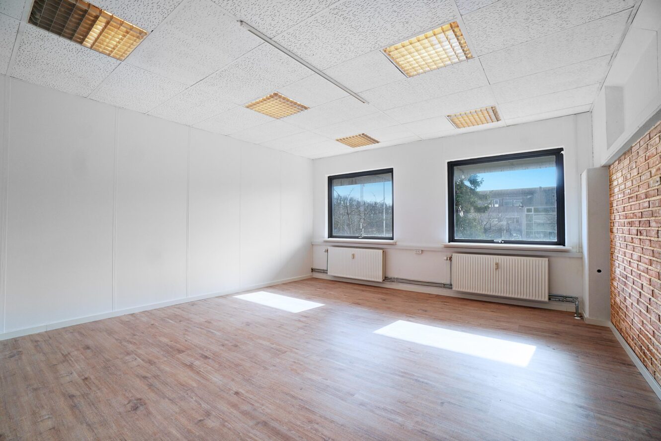 Tomt kontorlokale med trægulv, hvide vægge, to store vinduer, en murstensvæg og lysstofrør i loftet.