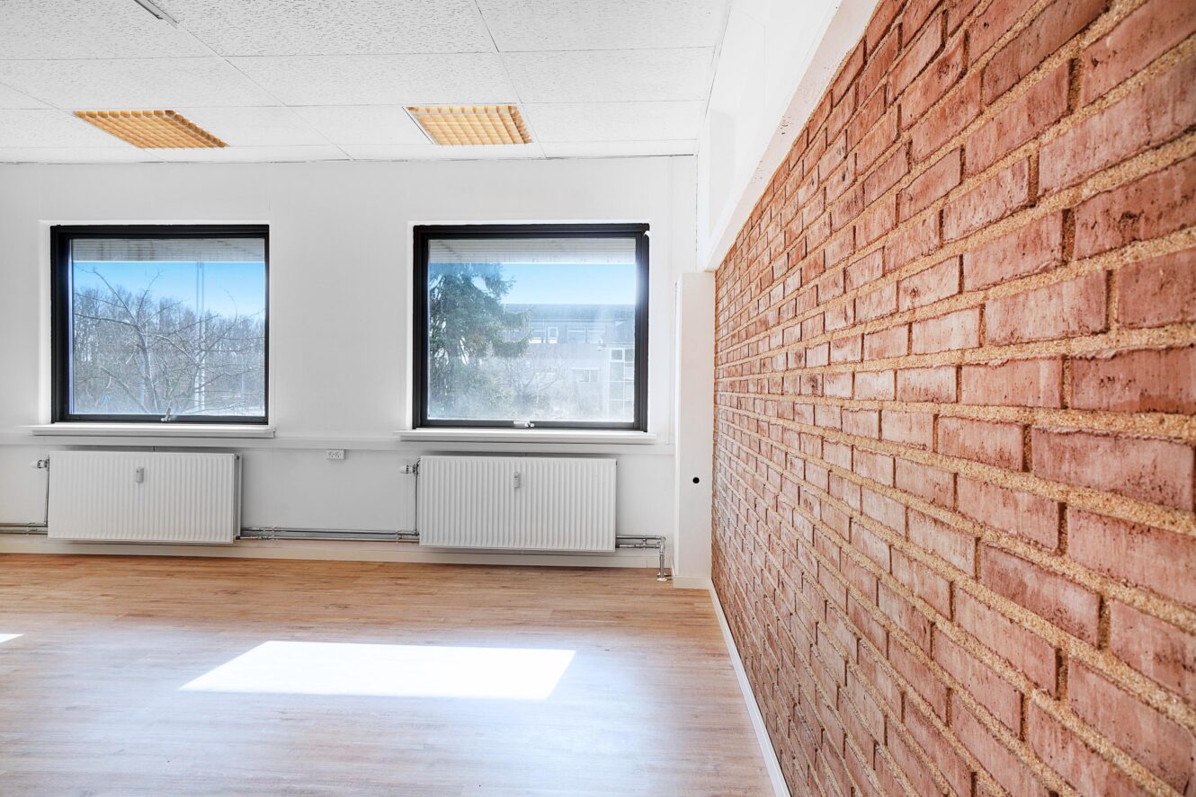 Et lyst, tomt rum med trægulv, hvide vægge, en rød murstensvæg med accent, to vinduer og to hvide radiatorer under vinduerne.
