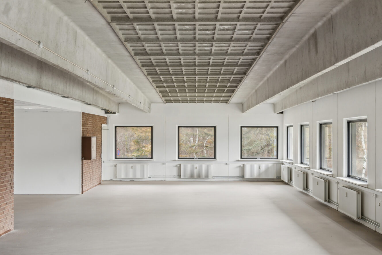 Rummeligt, tomt rum med store vinduer på to vægge, hvide vægge, eksponeret betonloft og en murstensvæg til venstre.