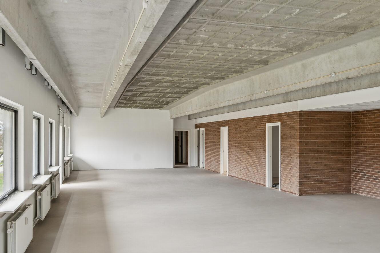Stort, tomt rum i industristil med betongulv, synligt betonloft, muret indervæg, flere døre og flere vinduer, der lukker naturligt lys ind.