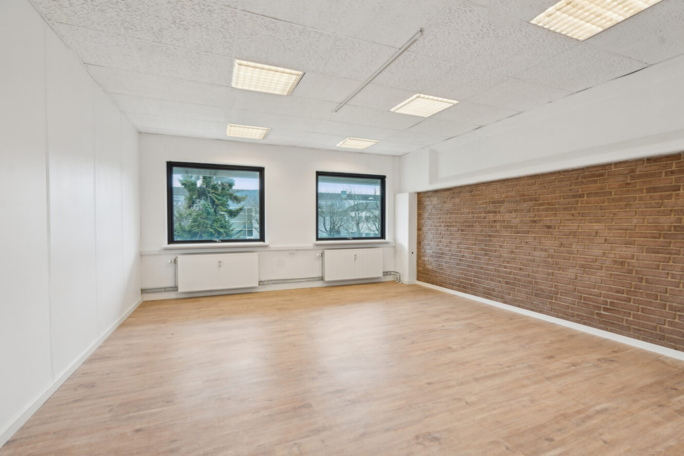 Tomt kontorlokale med trægulv, hvide vægge, en brun murstensvæg, to store vinduer og loftslamper.