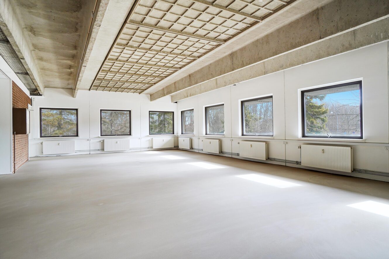 Tomt, rummeligt lokale med store vinduer på to vægge, hvide vægge, synligt betonloft og lyst gulv. Sollyset strømmer ind gennem vinduerne og skaber lyse pletter på gulvet.