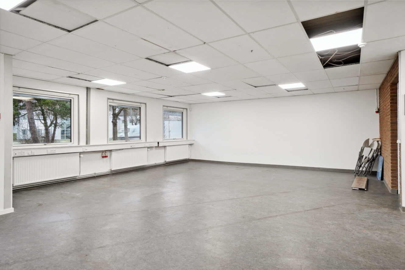 Tomt rum med hvide vægge, vinduer langs den ene side, en murstensvæg til højre, flere manglende loftsplader og stablede stole i det bageste hjørne.