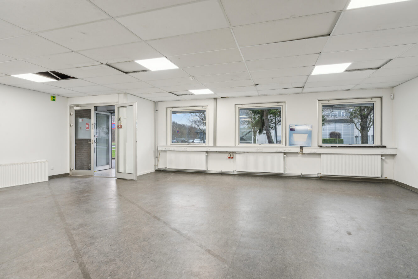 Et tomt rum med tre vinduer, hvide vægge, et gråt gulv, loftsplader og en åben dør, der fører ud. Nogle loftsplader mangler eller er forskudt.