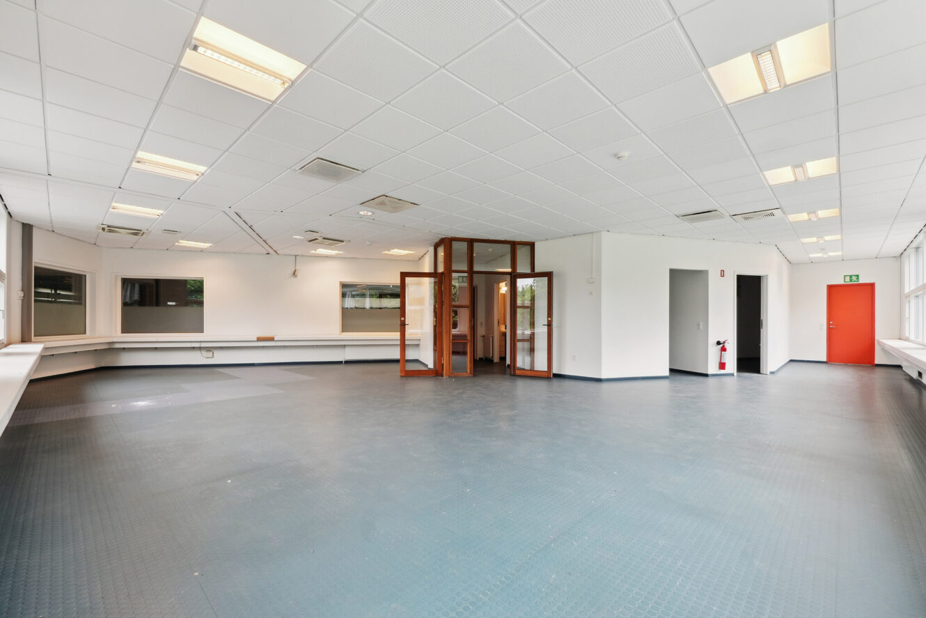 Rummeligt, tomt kontorlokale med store vinduer, hvide vægge, flisebelagt loft, gråt gulv og et lille kontorområde med glasvægge i midten.