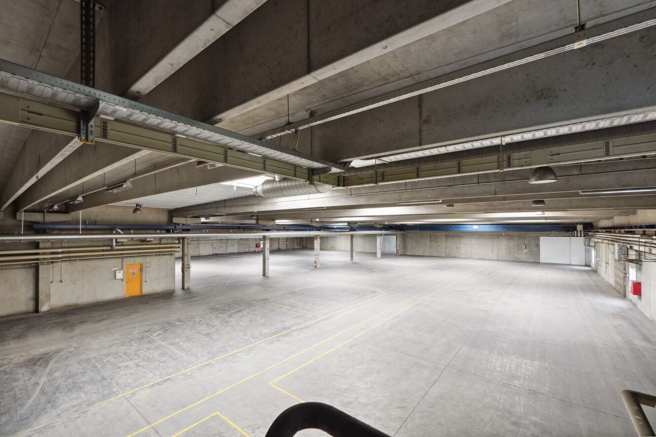 Bredt billede af en tom, ren, underjordisk parkeringskælder i beton med markerede parkeringspladser, industriel belysning og synlige rør i loftet.