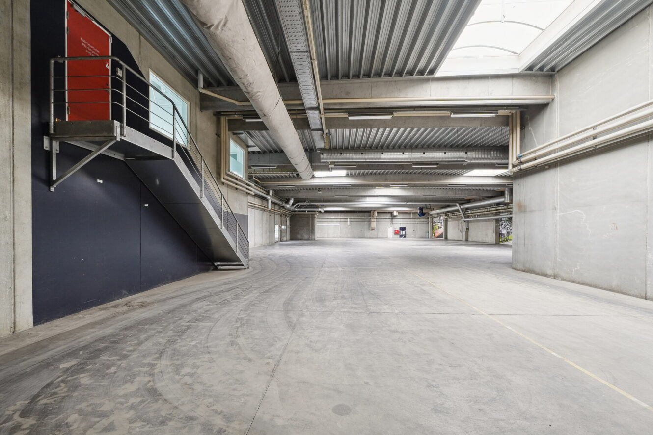 Stort, tomt industrilager med betongulv, højt til loftet, synlige rør og en metaltrappe, der fører op til en rød dør på en øvre platform. Naturligt lys kommer ind gennem ovenlysvinduer.