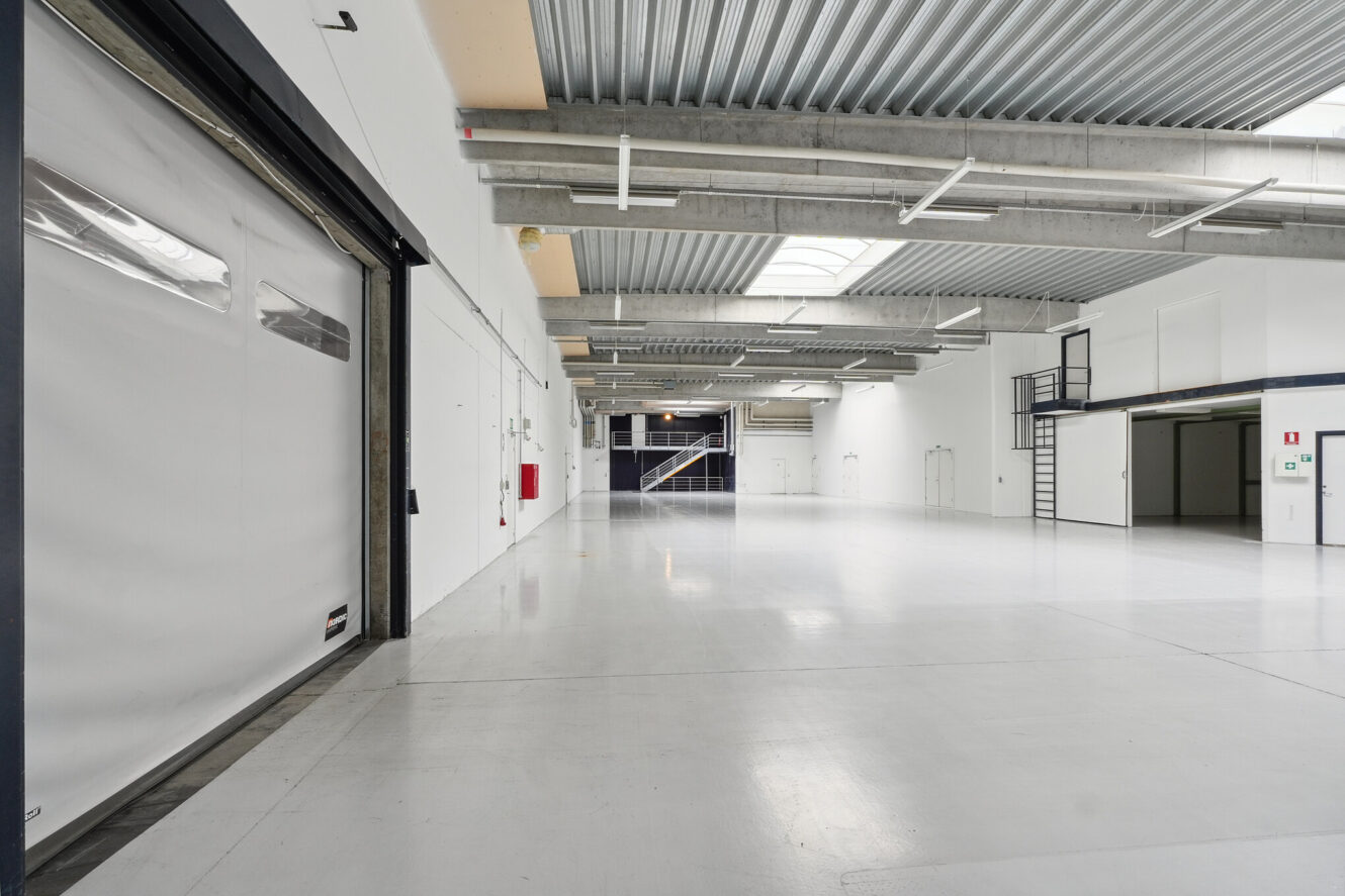 Stort, tomt industrilager med hvide vægge, store ovenlys, betongulv, åben mezzanin og synlige konstruktionsbjælker.