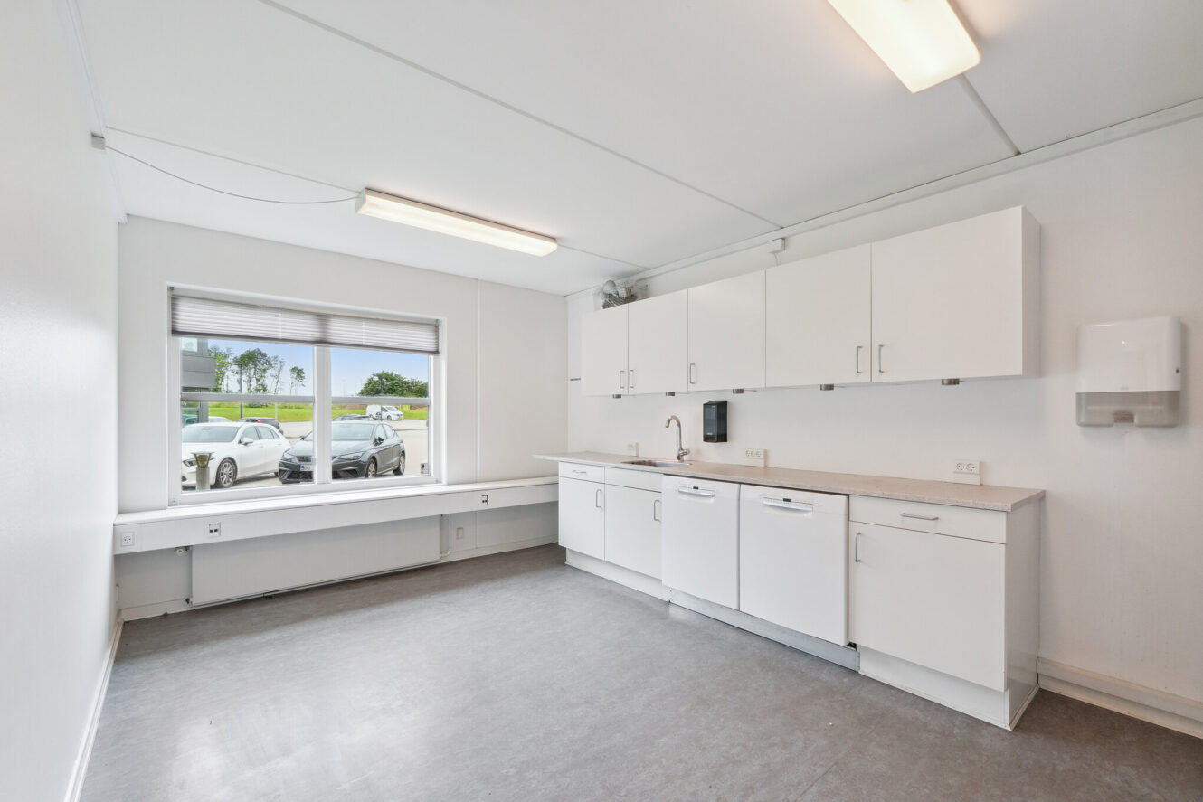 Et rent, moderne tekøkken med hvide skabe, bordplade og vask, stort vindue med udsigt til det fri og kraftig ovenlys.