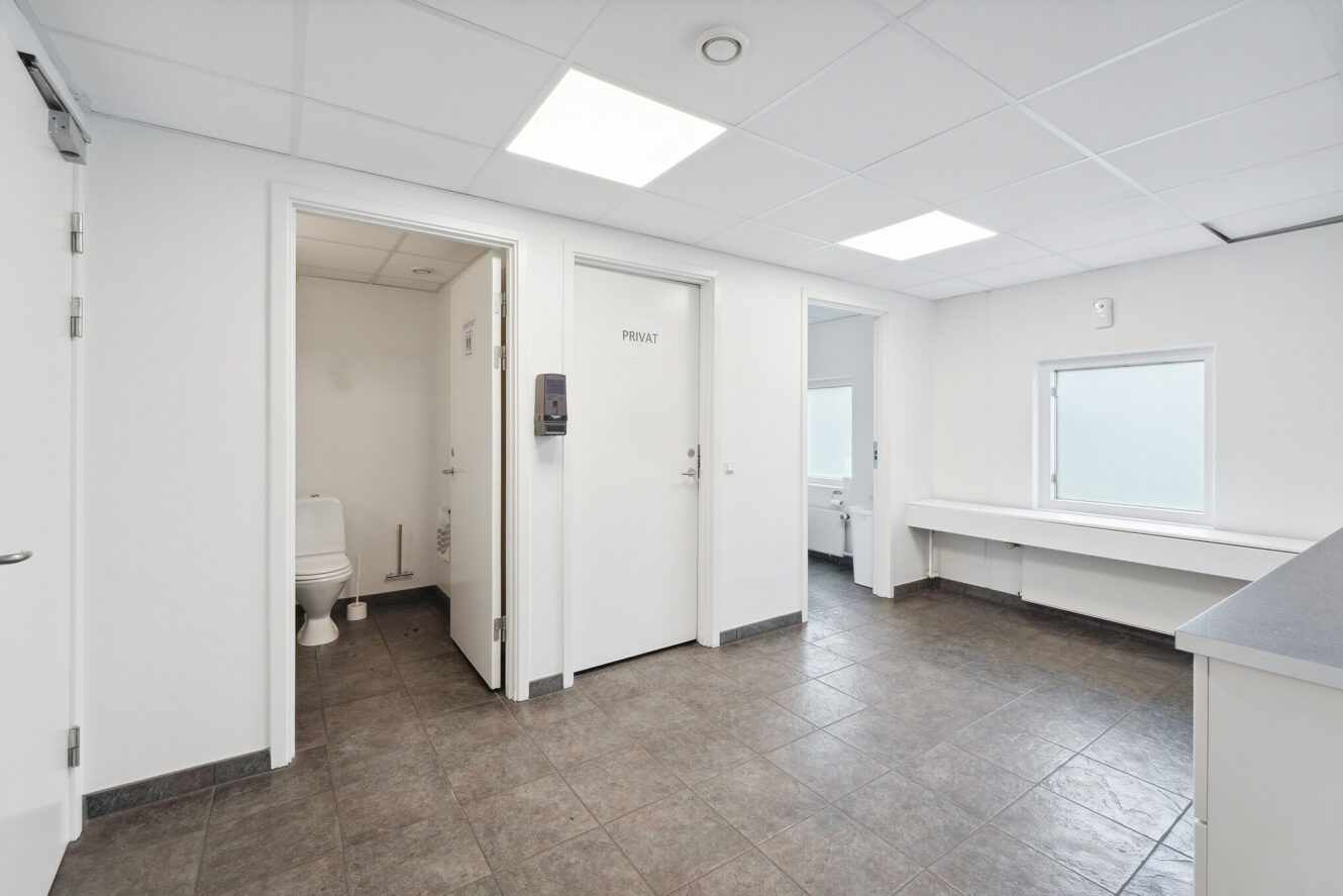 Et rent, moderne toiletområde med to hvide døre, hvoraf den ene står lidt åben og viser et toilet, klinkegulv og et vindue, der lukker naturligt lys ind.