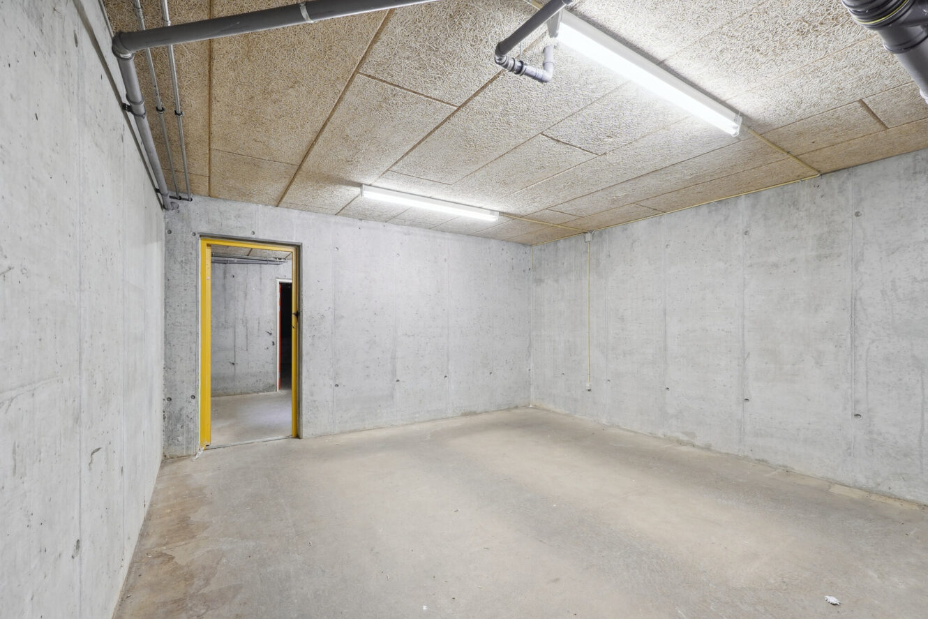 Umøbleret kælderrum med betonvægge, -loft og -gulv, synlige rør, lysstofrør og en åben døråbning med gul karm.