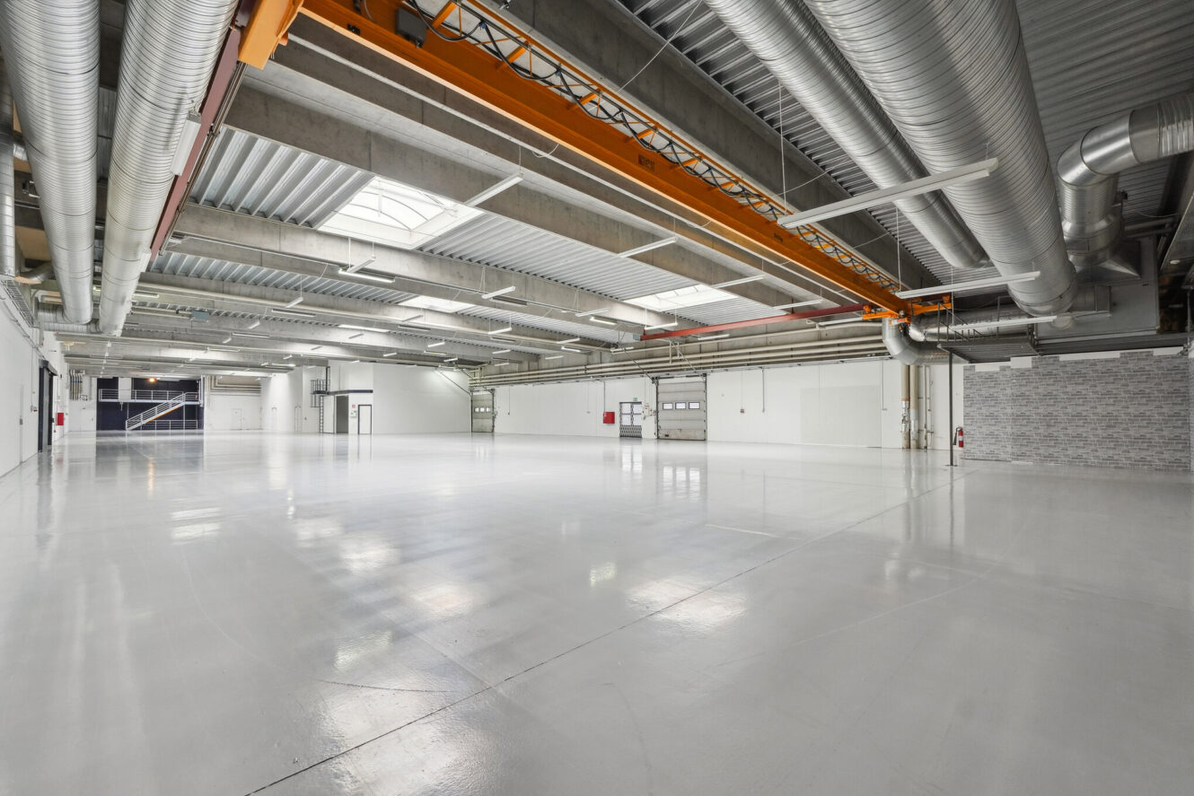 Stort, tomt industrilager med polerede gulve, synlige kanaler, stor traverskran og hvide vægge, oplyst af ovenlysvinduer og lysstofrør.