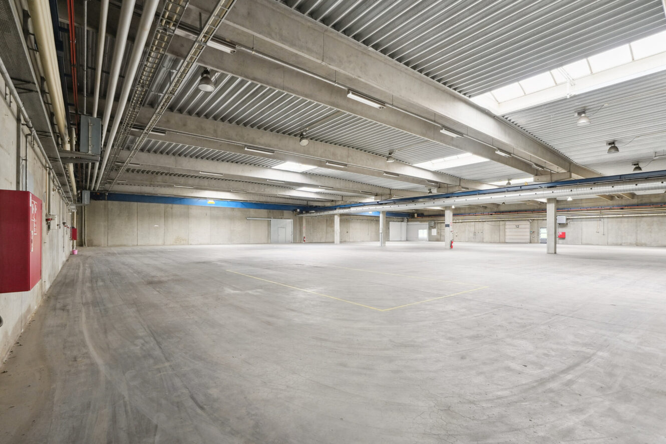 Stort, tomt industrilager med betongulv, højt til loftet, ovenlysvinduer, synlige rør og brandsikkerhedsudstyr på væggene.