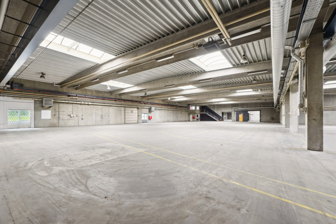 Tomt industrielt lagerinteriør med betongulv, højt til loftet, synlige kanaler og ovenlys. Gule linjer markerer gulvet; døre og trapper er synlige i den fjerneste ende.