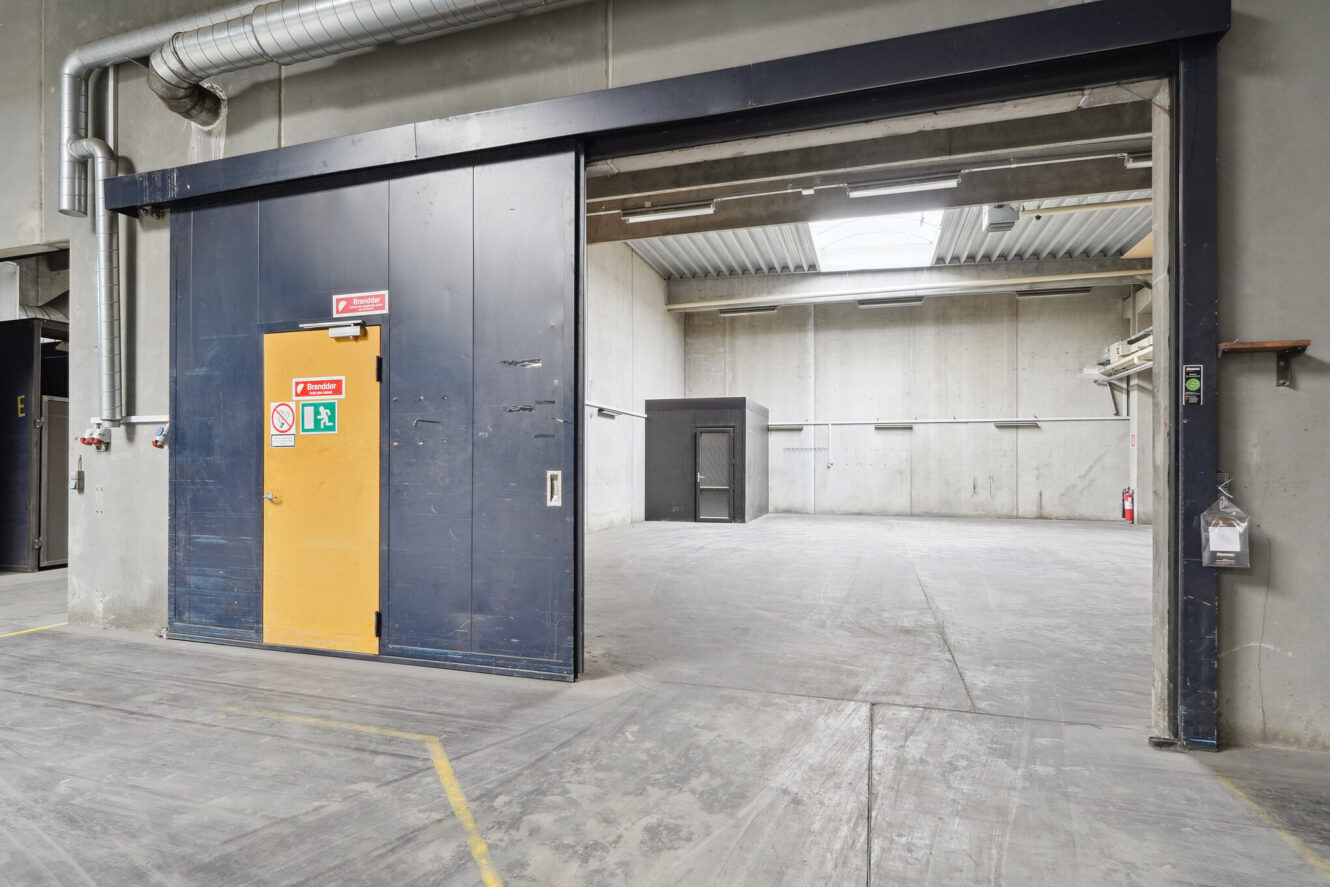Stort industrielt lagerinteriør med betonvægge og -gulve, skydedør i metal, ventilationsrør og synlige sikkerhedsskilte. Rummet virker tomt og godt oplyst.