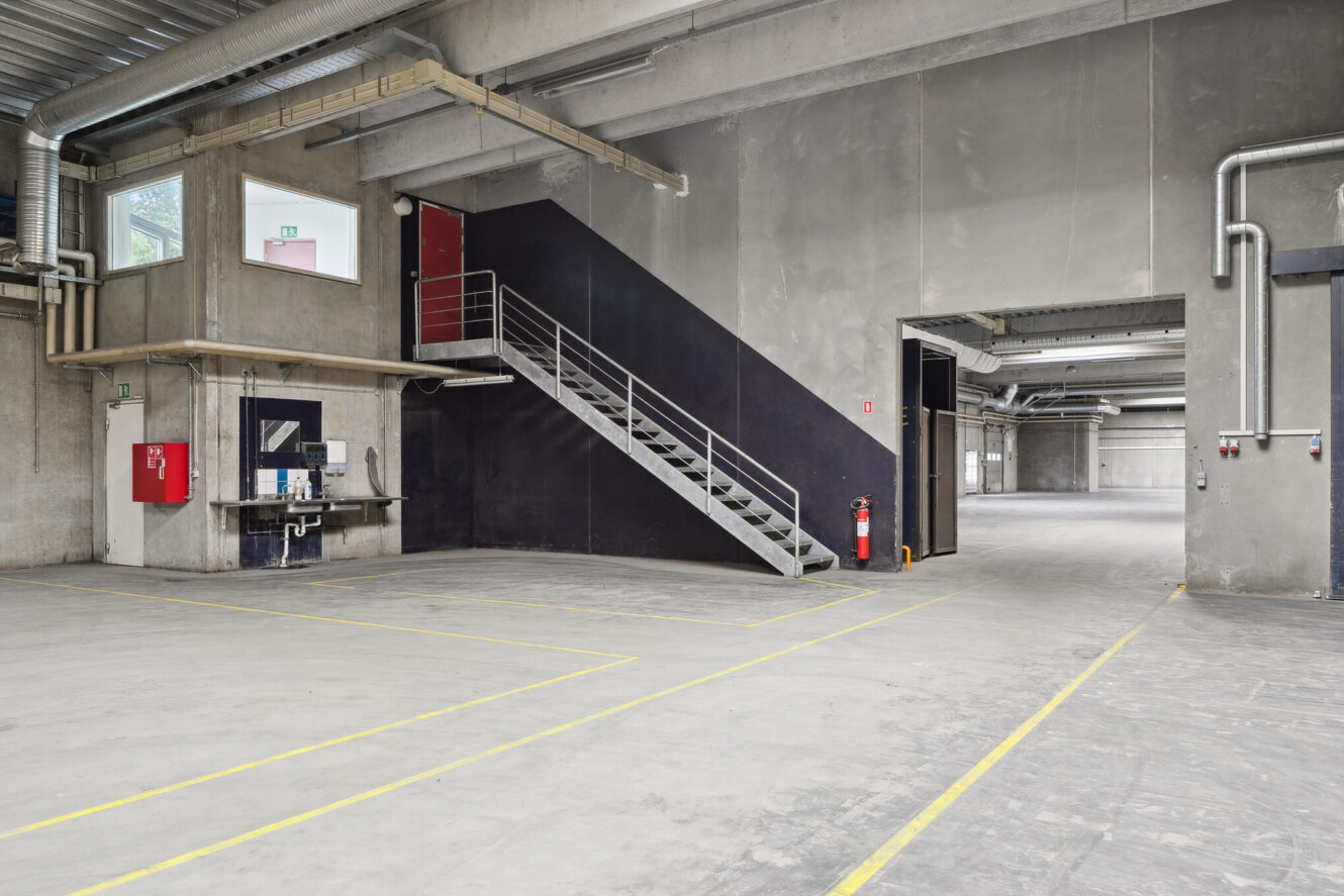 Interiør af et tomt industrilager med betongulve og -vægge, metaltrapper, sikkerhedsudstyr og gule streger, der markerer gulvet.