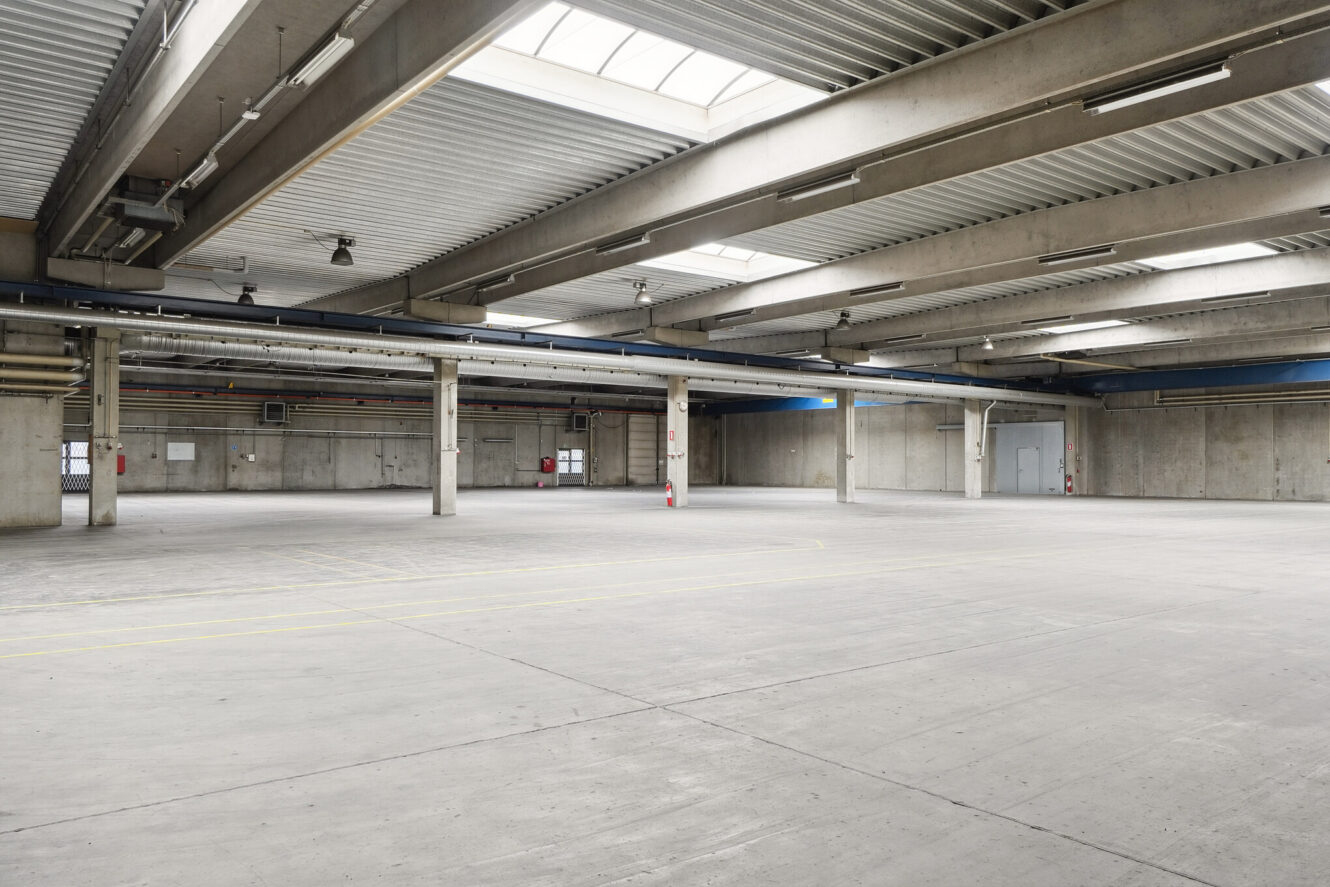 Stort tomt industrilager med betongulv, synlige bjælker, ovenlys og grå vægge; flere støttesøjler og brandslukkere er synlige.