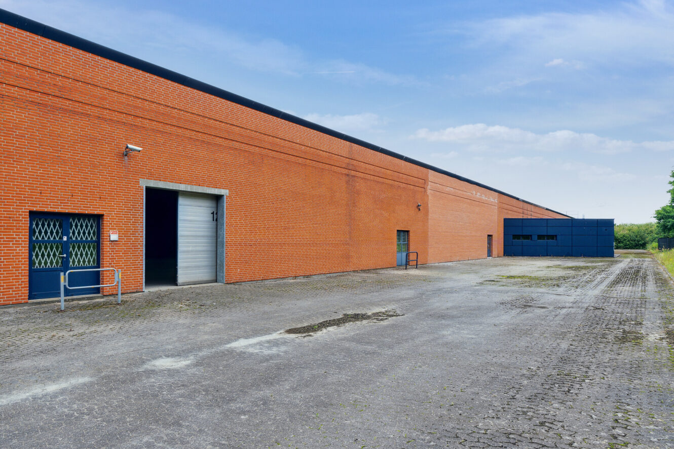 Lang muret industribygning med en stor garageport, sikkerhedsgitre i et vindue og et tomt asfalteret område foran under en delvis overskyet himmel.