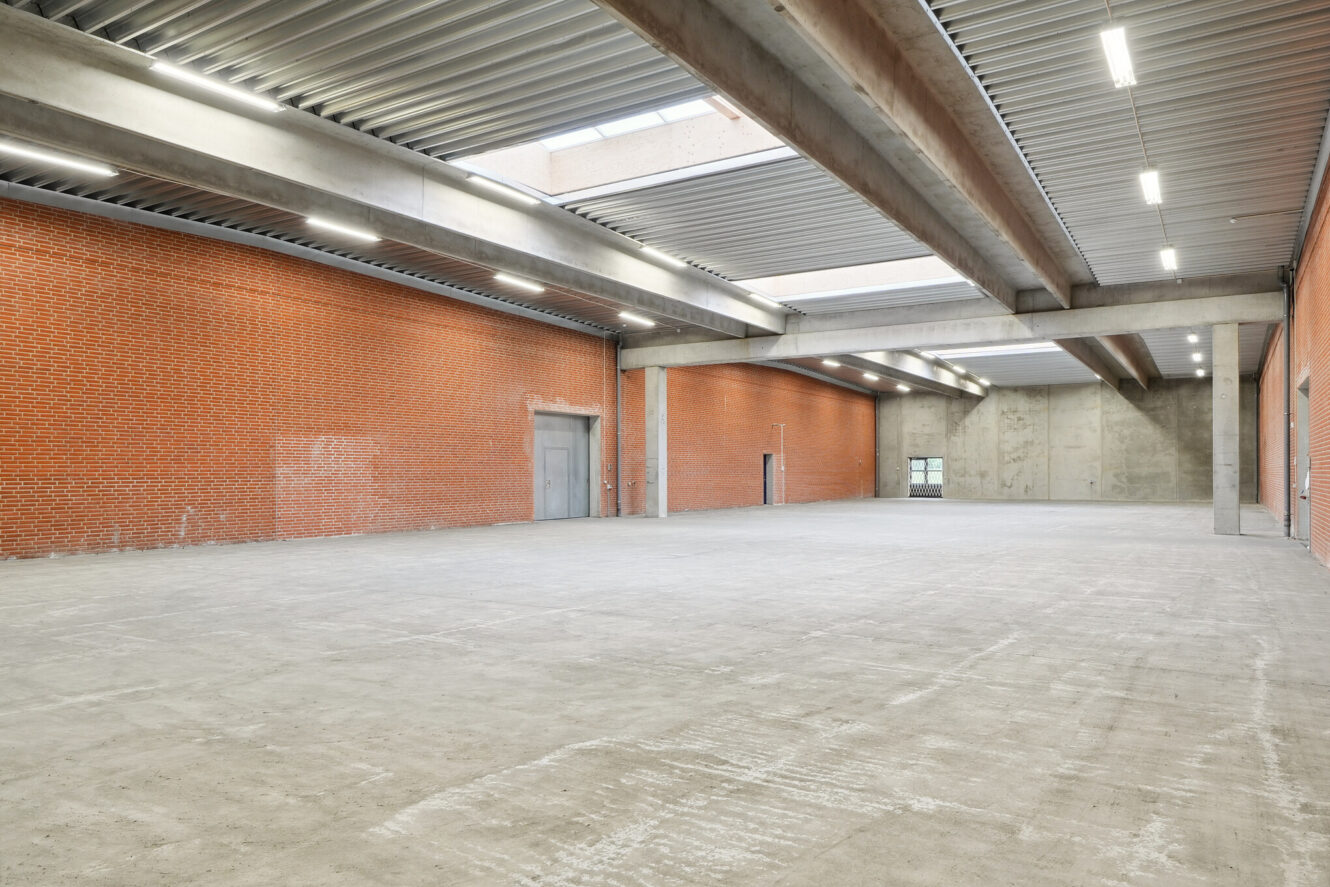 Stort tomt industrilager med betongulv, røde murstens- og betonvægge, synlige bjælker og lysstofrør i loftet.