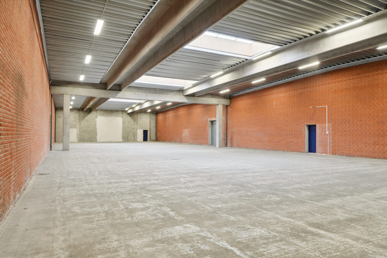 Stort, tomt industrilager med røde murstensvægge, betongulv, synlige loftsbjælker og fluorescerende belysning. Blå dør og hvid forsyningskasse på højre væg.