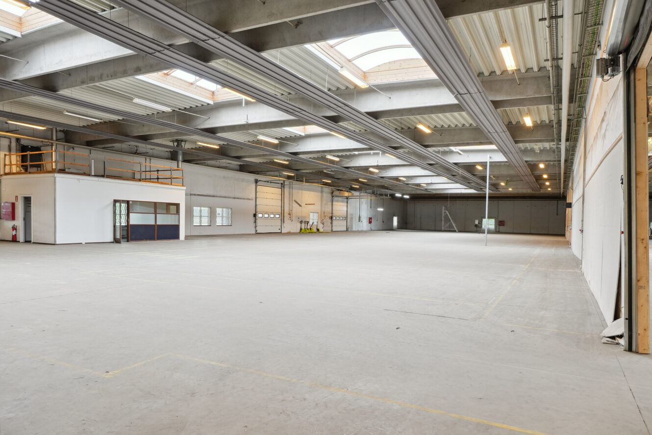 Rummelig tom lagerhal med betongulv, højt til loftet, ovenlys, lysstofrør og en øvre mezzanin i venstre side.