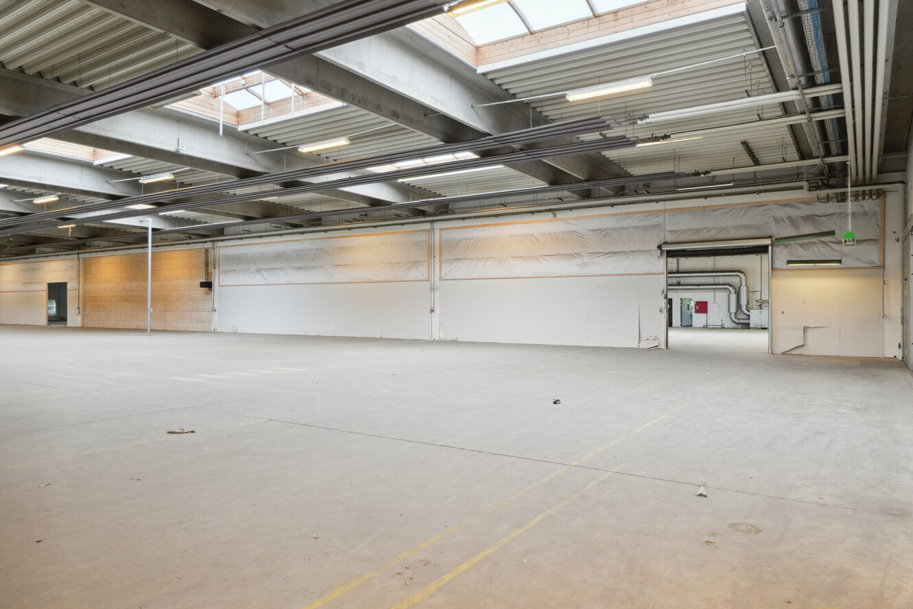 Stort tomt lagerinteriør med betongulv, synlige bjælker, ovenlys og hvide vægge; rør og en rød dør er synlige i baggrunden.
