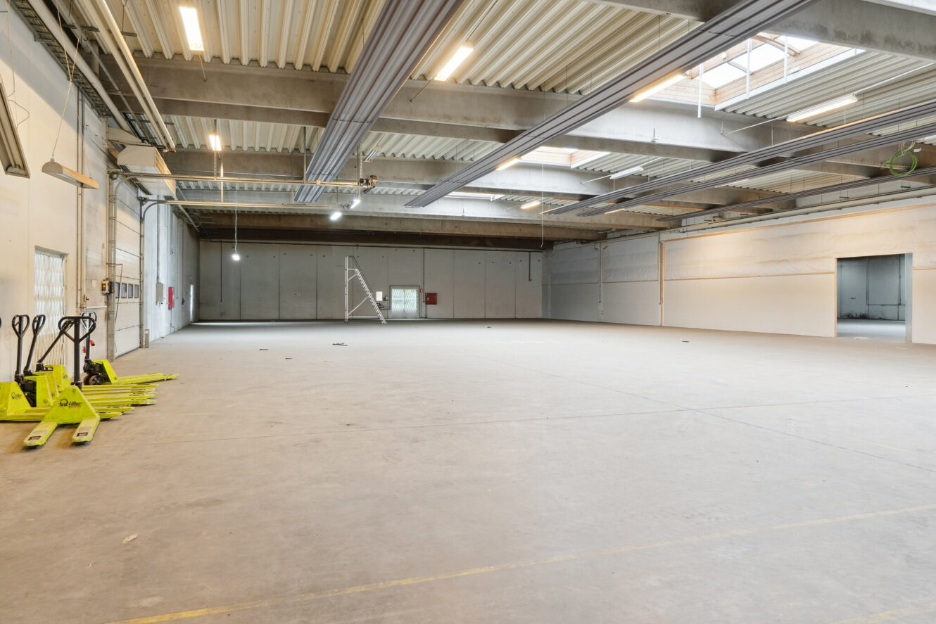 Stort, tomt lagerinteriør med betongulv, højt til loftet, synlige bjælker, ovenlys, palleløftere til venstre og en metalstige nær bagvæggen.