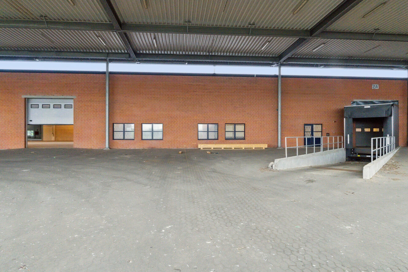 En læsserampe med en hævet rampe, en lukket garageport og flere små vinduer i en murstensbygning under et metaltag.