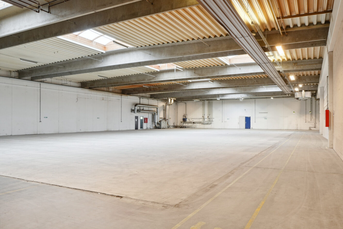 Rummeligt, tomt industrilager med betongulve, højt til loftet, synlige bjælker og store ovenlysvinduer, der giver naturligt lys indenfor.