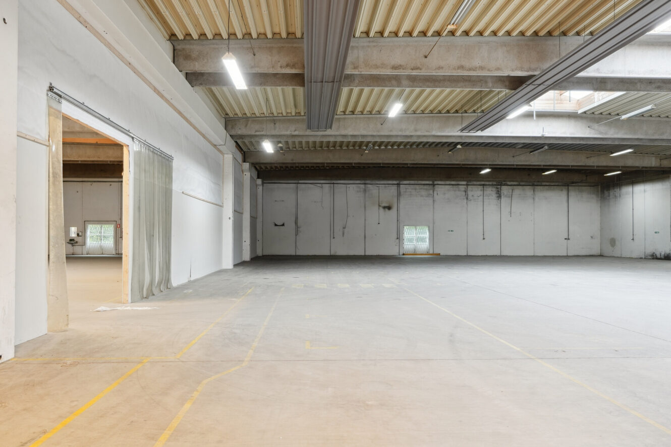 Stort, tomt industrilager med betongulv, højt til loftet, synlige bjælker, fluorescerende belysning og minimale vinduer. Et plastikgardin dækker delvist en åben døråbning til venstre.
