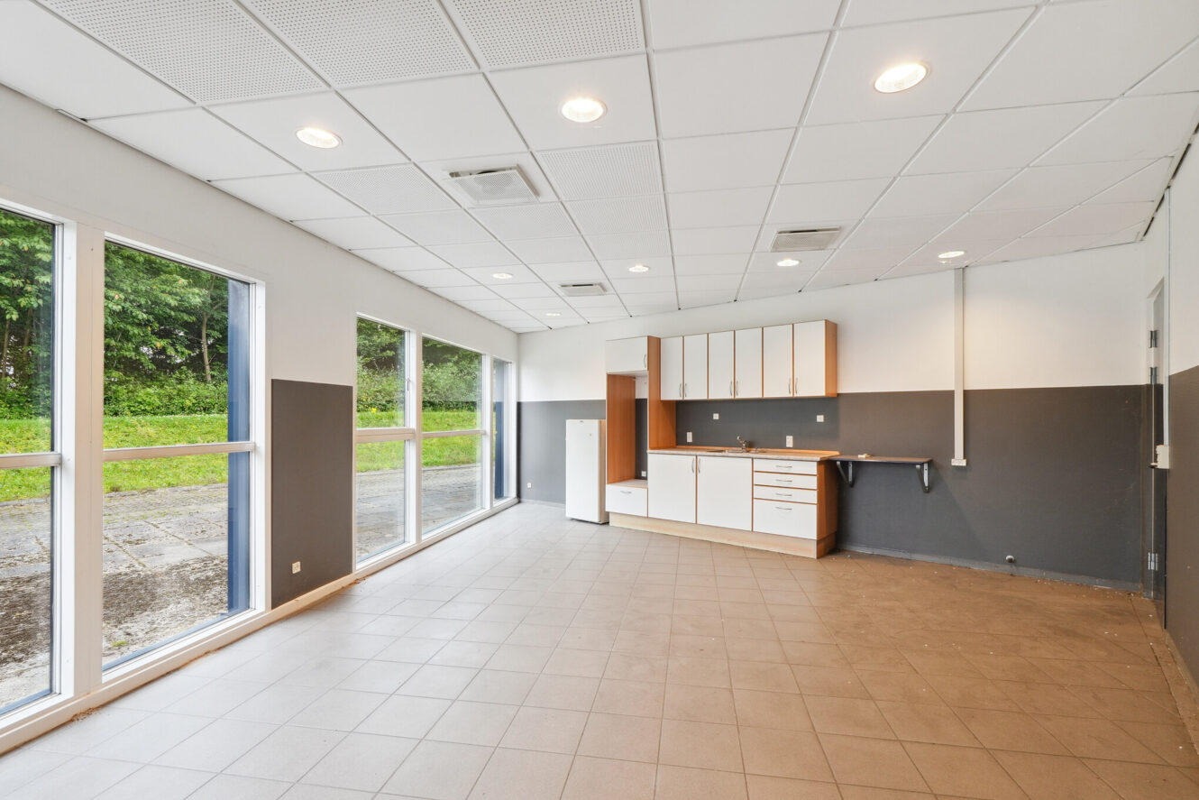 Stort, tomt rum med klinkegulv, loftslamper, indbyggede skabe, køleskab og vinduer langs den ene væg, der viser et græsklædt udendørsareal.