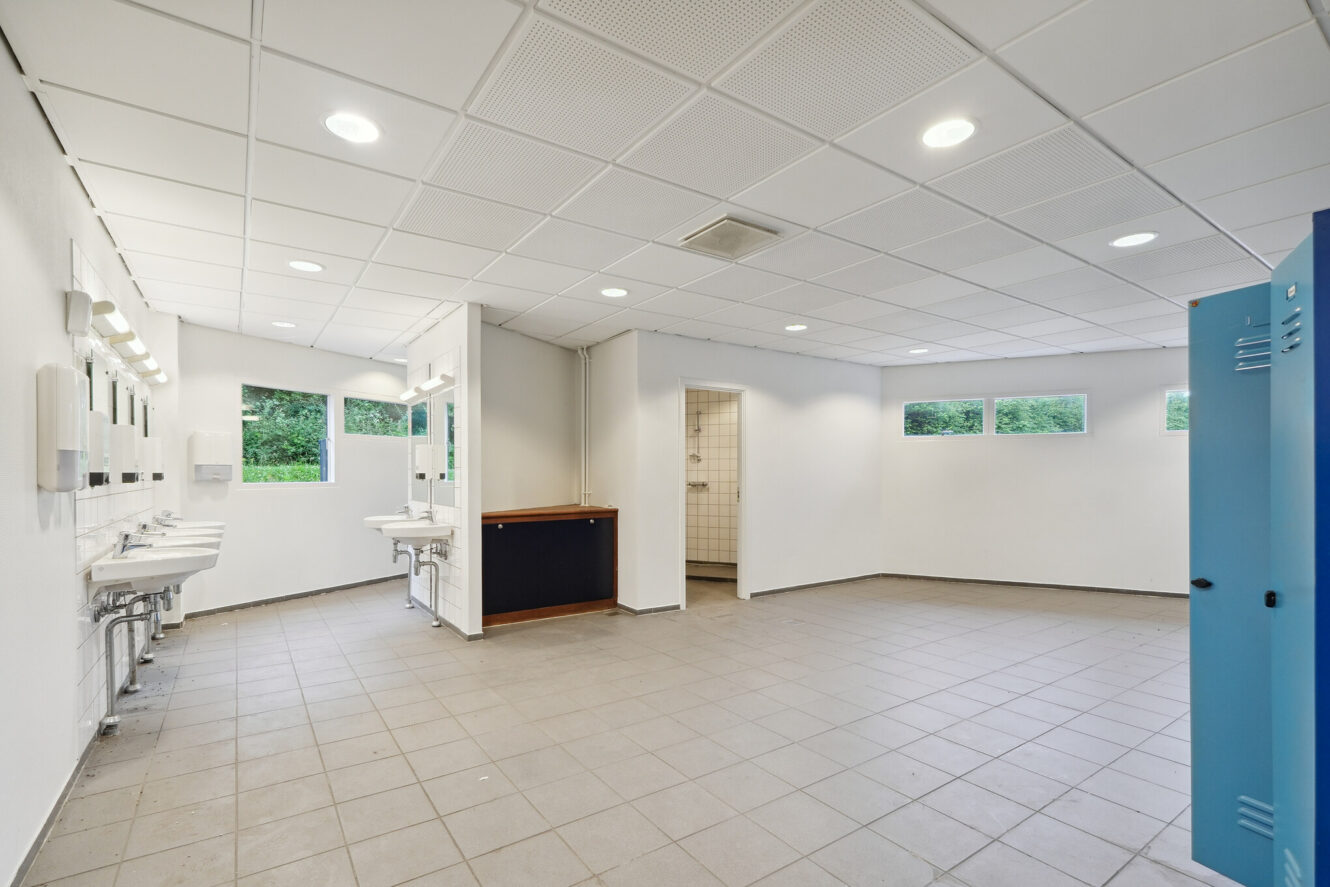 Rummeligt offentligt toilet med klinkegulv, flere håndvaske med spejle, blå skabe til højre og naturligt lys fra flere vinduer.