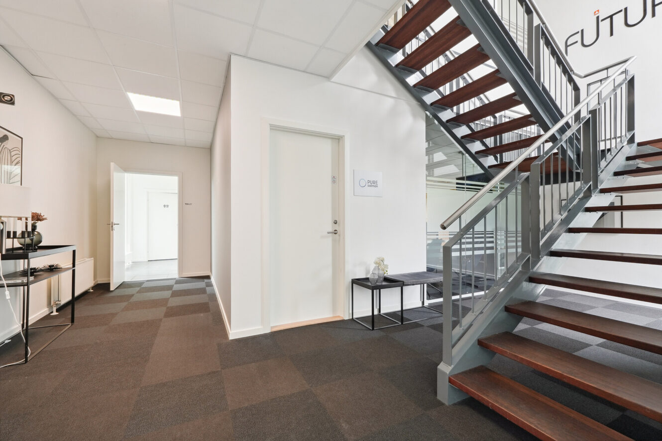 Moderne kontorgang med en trappe, en lukket hvid dør, en bænk og minimal indretning; Futur delvist synlig på væggen over trappen.