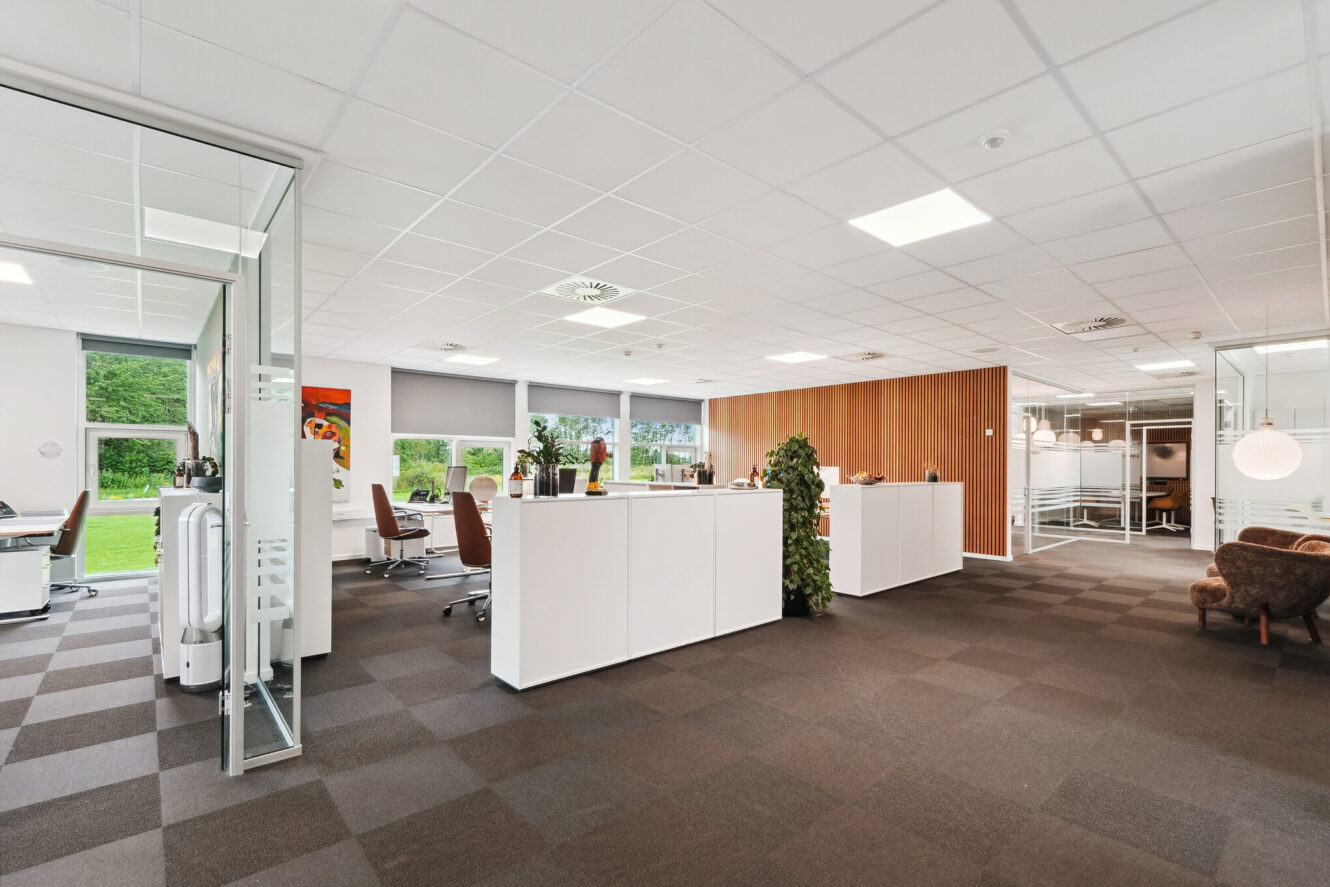 Moderne åbent kontorlandskab med hvide skriveborde, brune stole, glasvægge, indendørs planter og store vinduer, der lukker naturligt lys ind.