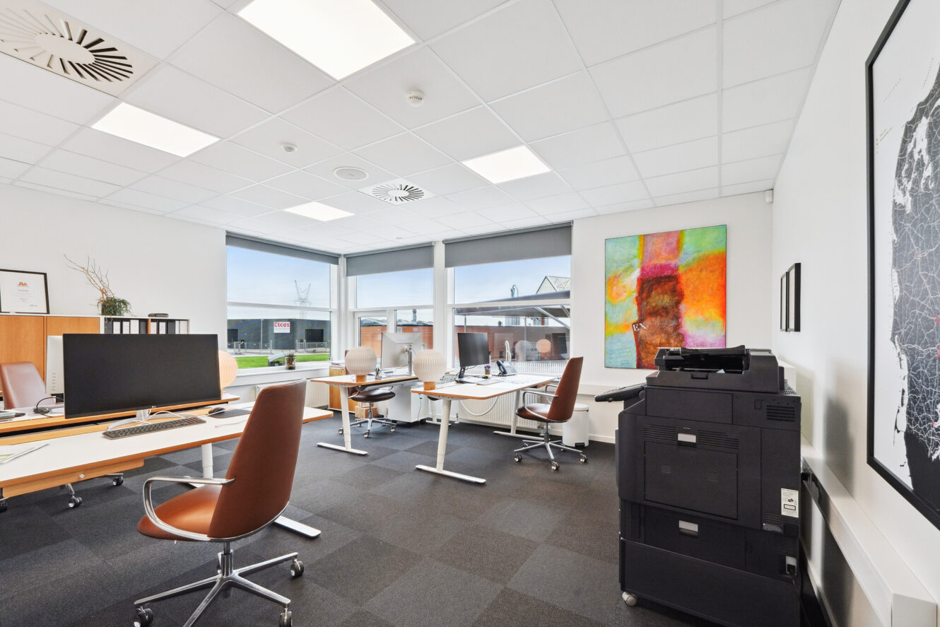 Moderne kontor med brune stole, skriveborde, computere, en printer, store vinduer og farverige kunstværker på en hvid væg.