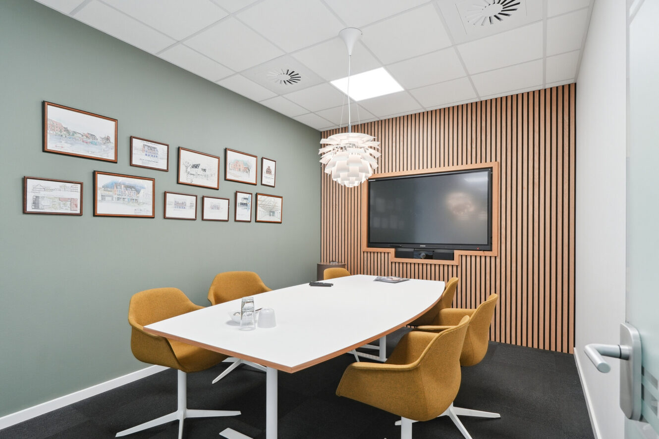 Moderne konferencelokale med et hvidt bord, seks sennepsgule stole, en stor skærm på en træbeklædt væg, pendellamper og indrammede billeder på en grøn accentvæg.