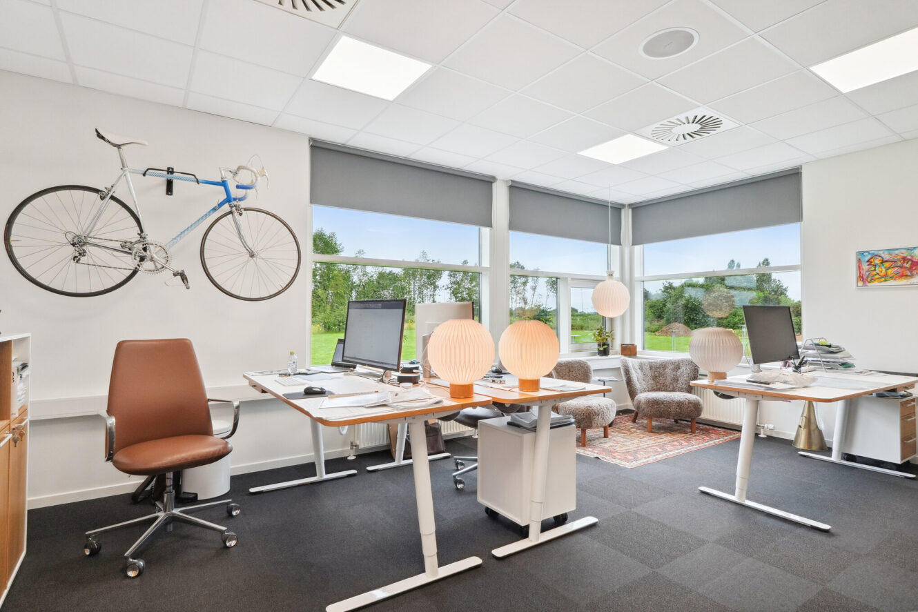 Moderne kontor med store vinduer, to skriveborde med computere, brune stole, hængende cykel på væggen, papirlamper, en lænestol og en grøn udendørs udsigt.