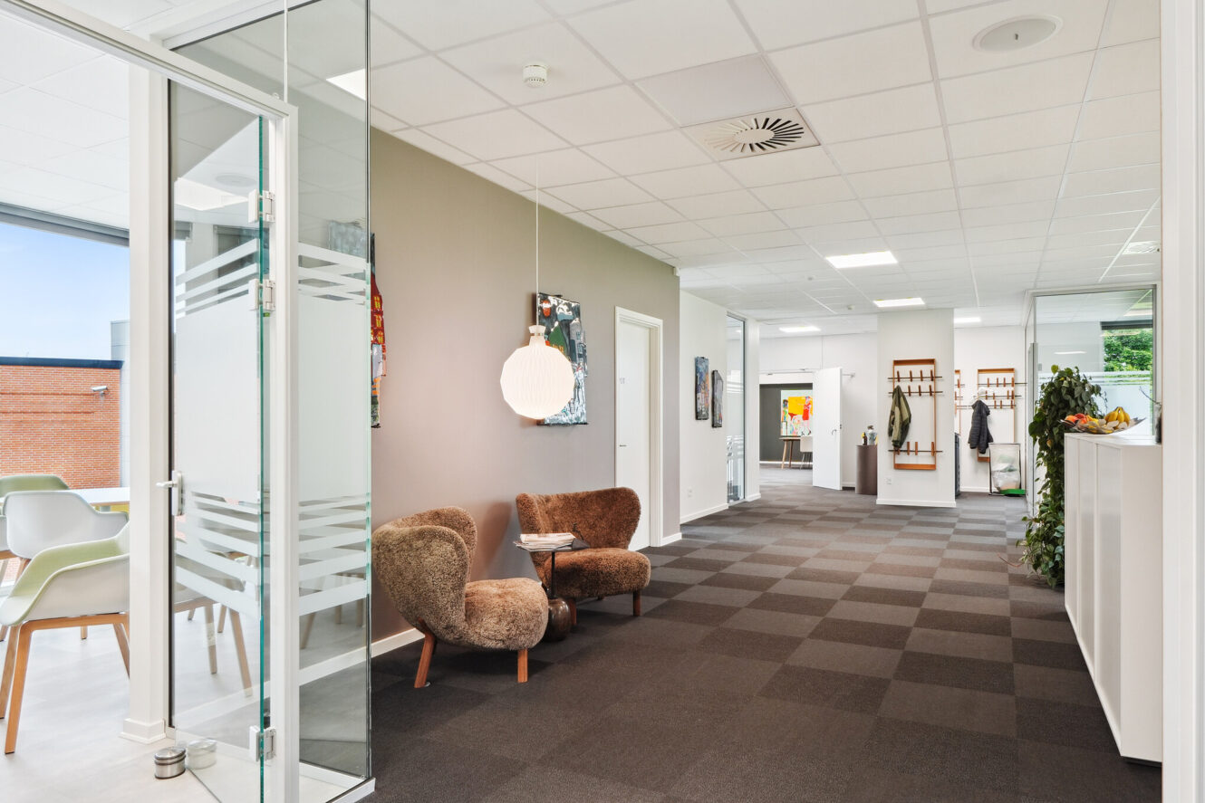 Moderne kontorgang med ternet tæppe, to brune stole og et lille bord, mødelokale i glas til venstre, knagerækker i den anden ende og planter langs et hvidt skab.
