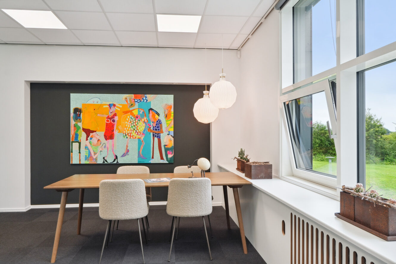 Et moderne mødelokale med et træbord, fire polstrede stole, farverige abstrakte kunstværker på væggen og store vinduer, der lukker naturligt lys ind.