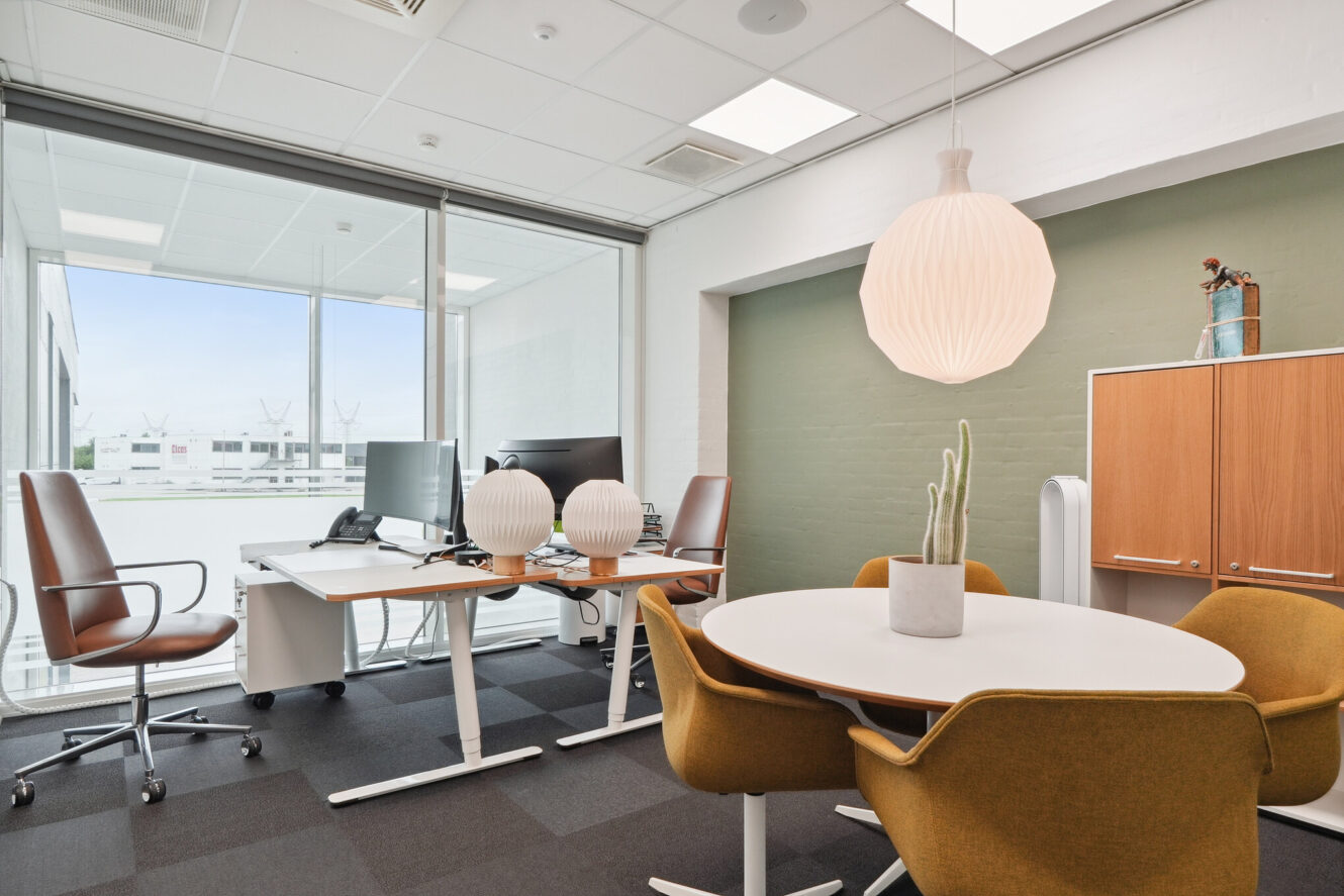 Moderne kontor med to skriveborde, computere og to brune stole nær et vindue; rundt bord med fire gule stole og en kaktus i potte; skab og pendellampe i baggrunden.