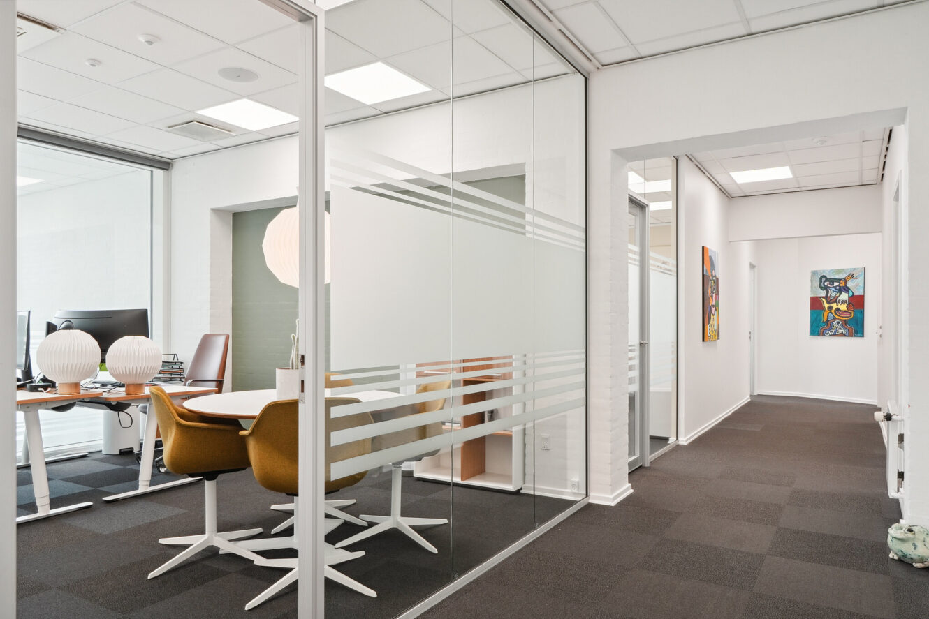 Moderne kontorgang med mødelokale med glasvægge, rundt bord, brune drejestole, computere og abstrakt kunst på hvide vægge.
